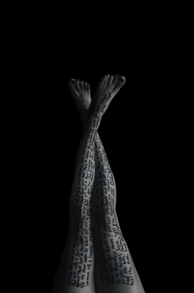 A pair of crossed legs, photographed from the knees down against a dark background. The legs are covered in handwritten text that runs down the length of the skin, with the words appearing in black ink. The stark contrast between the pale skin and the dark background emphasizes the flowing lines of the text.